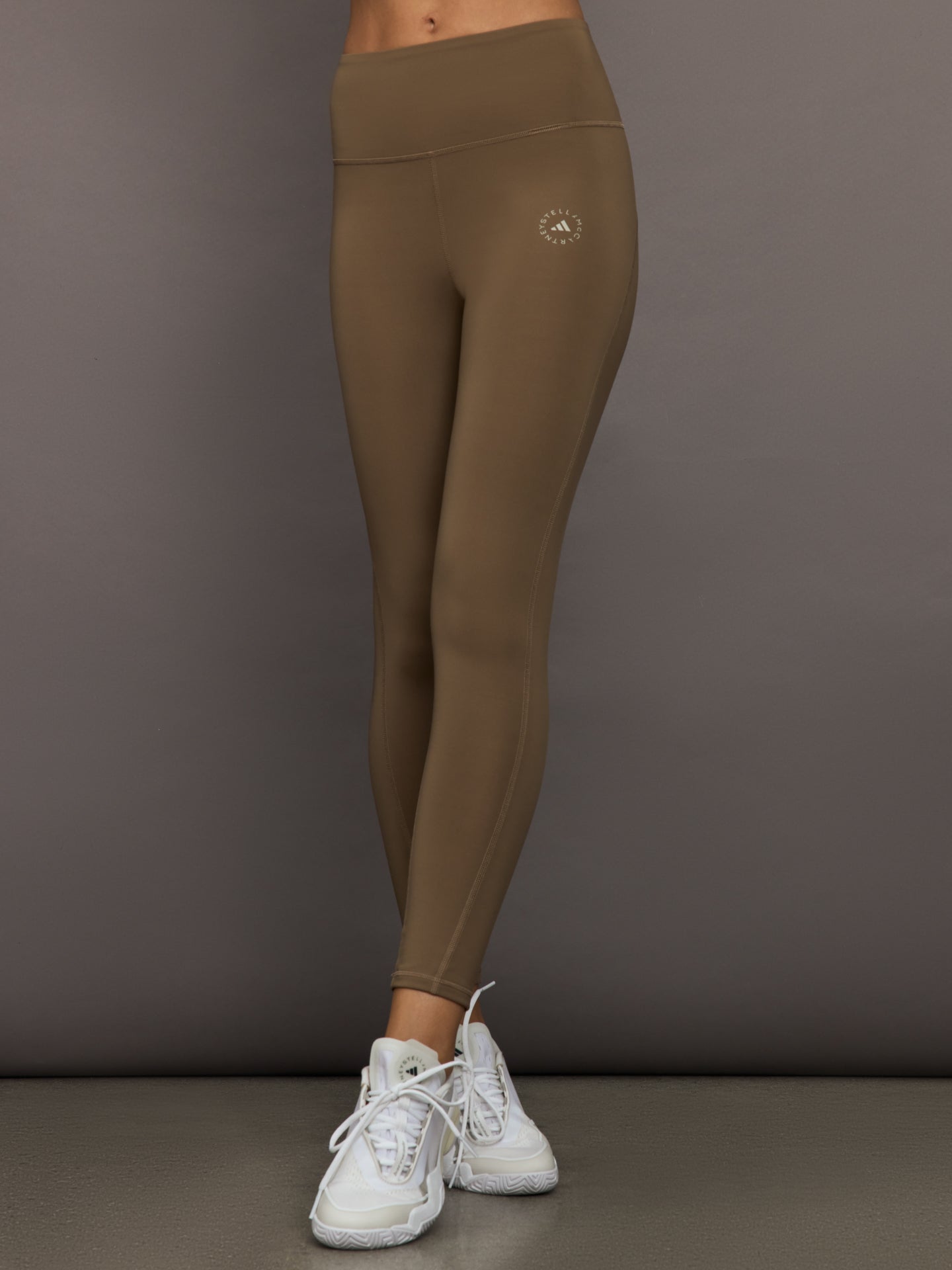 ADID-JW2001-BRWNBRWDRK-aSMC-TrueStrength-Yoga-7-8-Leggings-color-Brown-Moss_36988-Edit Person wearing brown leggings with a logo on a gray background
