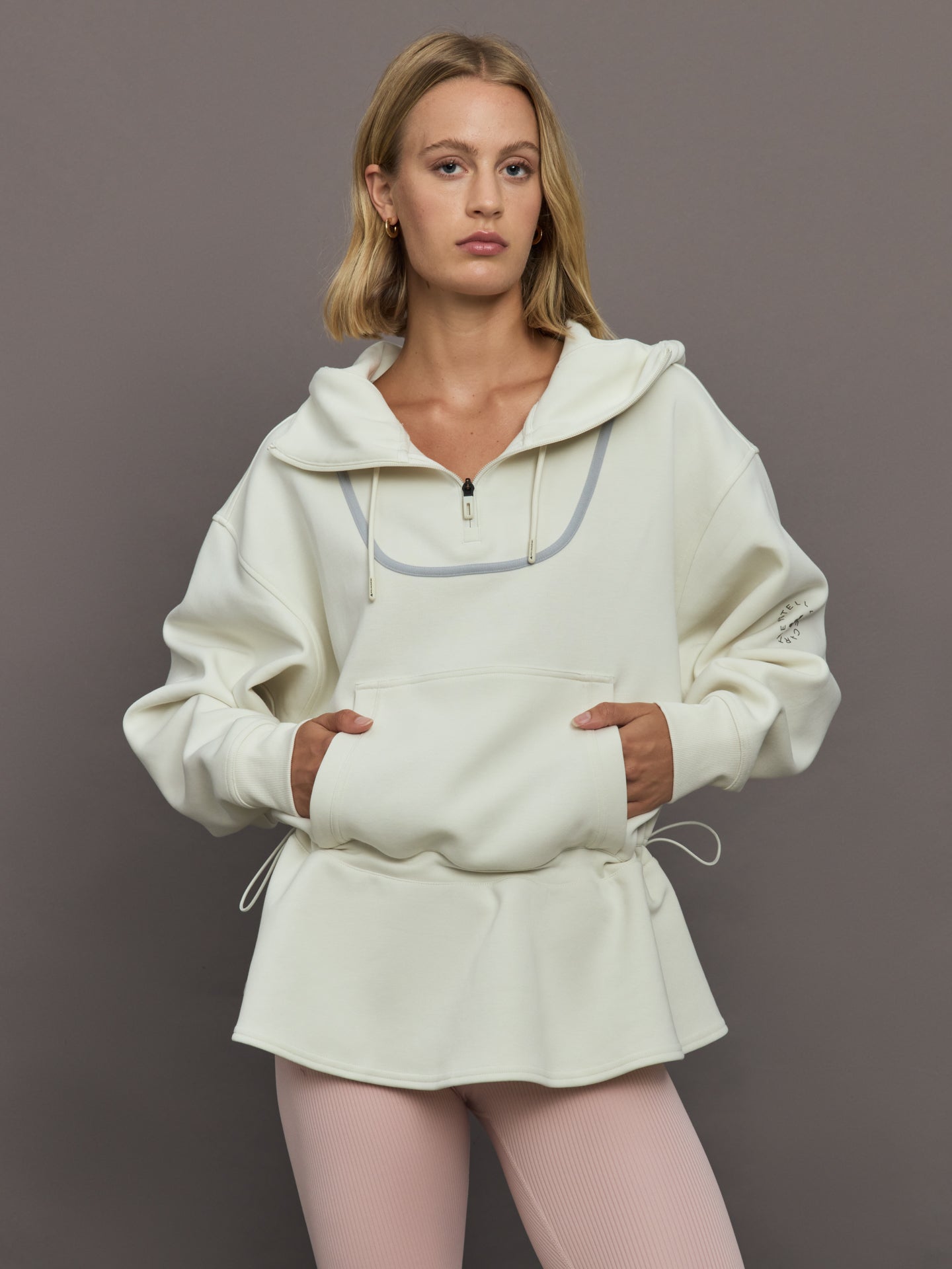 ADID-JX9108-IVRYOFFWHI-aSMC-Peplum-Hoodie-color-Almond-Milk-Smc_11570-Edit Model wearing a cream-colored hoodie with a quarter zip.