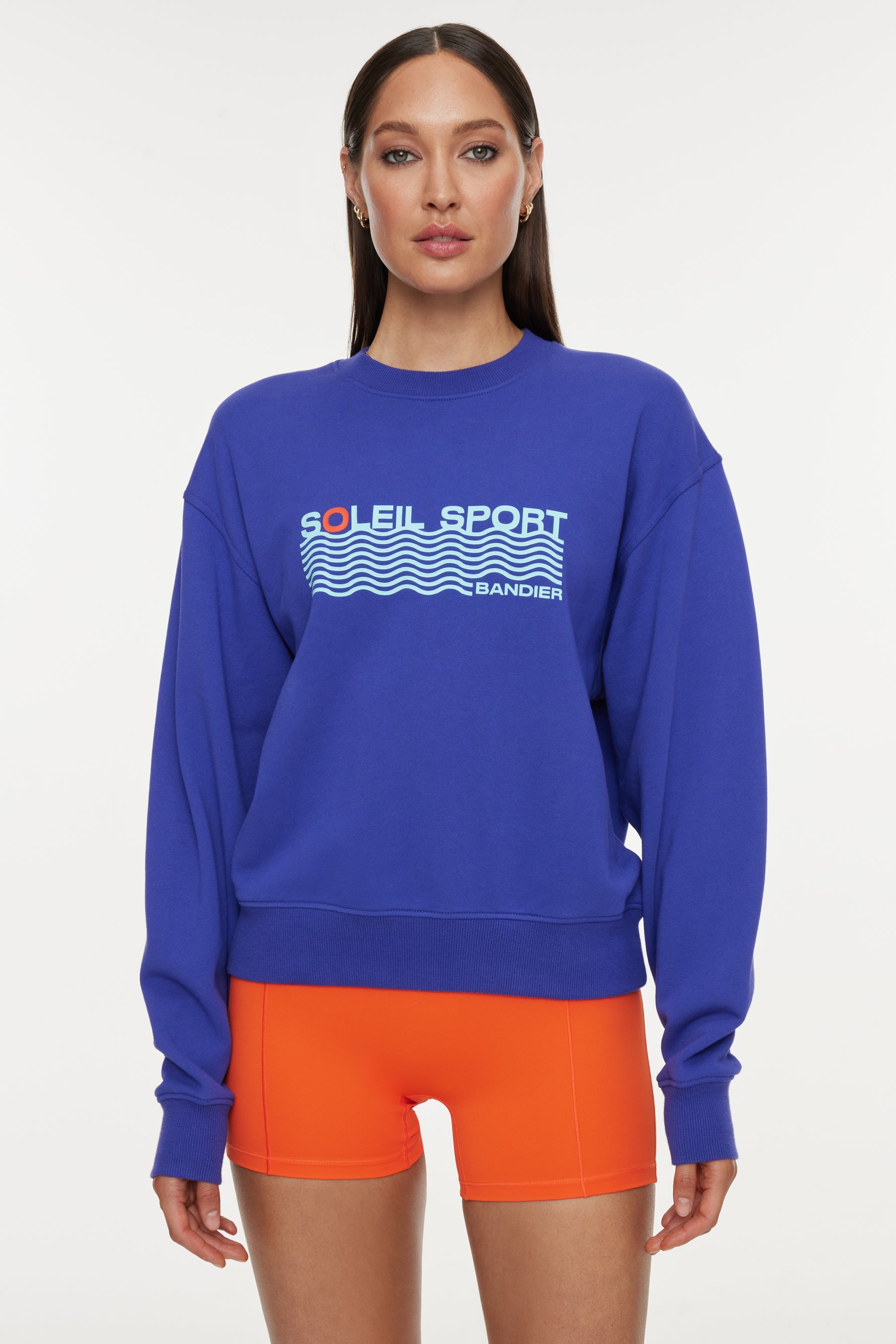 BND10425-SPECTRUMBLUE_1843-Edit_99e51caf-85f3-4507-a000-313438f0dc9f Model wears a royal blue crewneck sweatshirt with the lettering "Soleil Sport" across the chest with wavey lines below and "Bandier" in the bottom right corner.