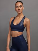 Model wears a navy, glossy, deep scoop-neck sports bra with a sleek racerback and a shiny, liquid-like finish.