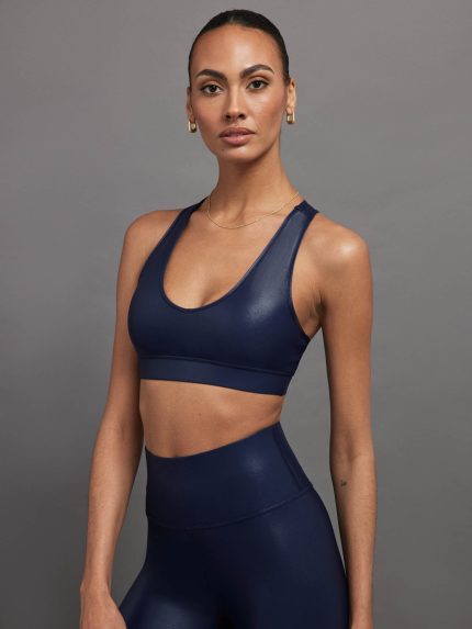 Model wears a navy, glossy, deep scoop-neck sports bra with a sleek racerback and a shiny, liquid-like finish.