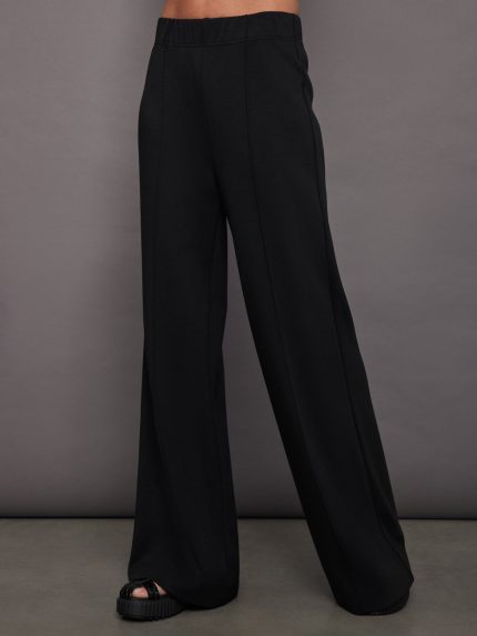 Model wears black relaxed-fit pants with a smooth finish and subtle pintuck seams down the front.