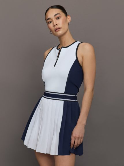 Model wears a sleeveless colorblocked white and navy high waisted tennis/golf dress with navy waistband and pleated skirt. 