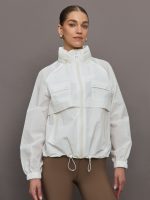 Model is wearing a white windbreaker with drawstrings at the bottom of the jacket and twin chested pockets and a detachable hood.