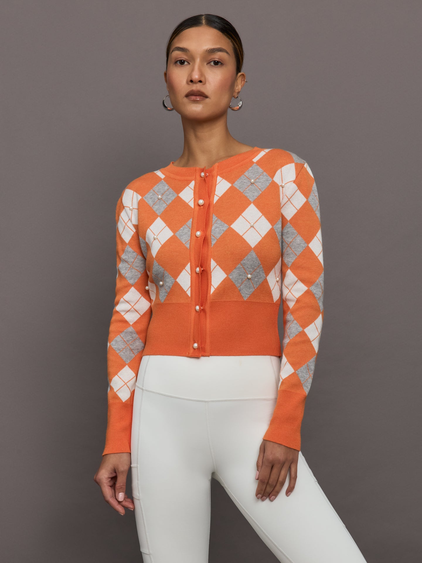 THEO-T2512K650-ORNGORNGMED-Nomia-Plaid-Pearly-Cardigan-color-Mandarine-Gold_10536-Edit Model wears an orange cardigan with white and gray argyle print and pearls centered over each argyle.